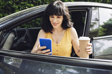 Glückliche Frau mit Einweg-Kaffeebecher und Smartphone beim Aussteigen aus einem Elektroauto - JCCMF03717