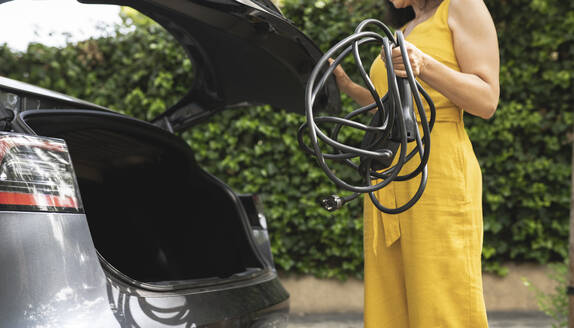 Woman holding cable while standing by open car trunk - JCCMF03729