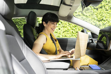 Glückliche Geschäftsfrau, die in einem Elektroauto sitzend einen Laptop benutzt - JCCMF03731