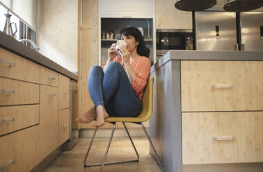 Mature woman drinking coffee while sitting on chair in kitchen at home - JCCMF03744