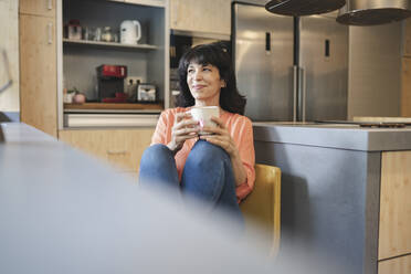 Lächelnde reife Frau mit Kaffeetasse sitzt in der Küche zu Hause - JCCMF03745