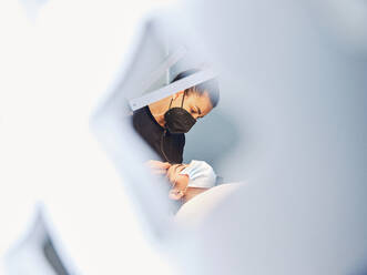 Through grate side view of professional cosmetician treating face of female client in protective mask during beauty procedure in salon - ADSF29279