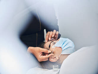 Anonyme Kosmetikerin mit Applikator, die die Augenbraue einer Kundin mit Vergangenheit für die Laminierung während einer Schönheitsbehandlung im Salon abdeckt - ADSF29281