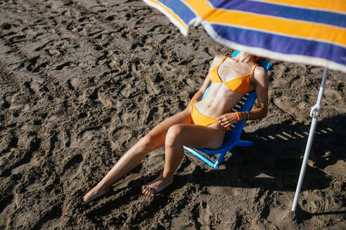 Anonyme junge rothaarige Frau sitzt auf einem Stuhl am Strand an einem sonnigen Tag im Sommer - ADSF29298