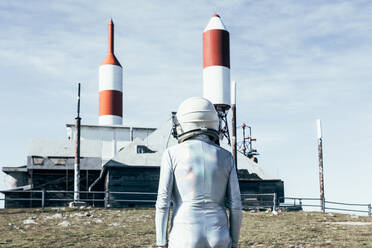 Back view man in spacesuit standing on rocky ground against metal fence and striped rocket shaped antennas on sunny day - ADSF29299