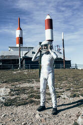 Full body man in spacesuit standing on rocky ground against metal fence and striped rocket shaped antennas on sunny day - ADSF29300
