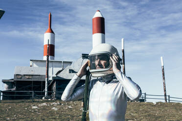 Mann im Raumanzug steht auf felsigem Boden vor einem Metallzaun und gestreiften raketenförmigen Antennen an einem sonnigen Tag - ADSF29301