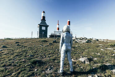 Back view full body man in spacesuit standing on rocky ground against metal fence and striped rocket shaped antennas on sunny day - ADSF29303