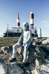 Ganzkörper-Mann im Raumanzug auf felsigem Boden sitzend gestreifte raketenförmige Antennen an einem sonnigen Tag - ADSF29304