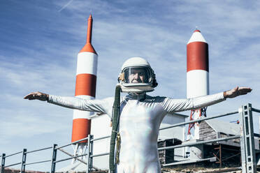 Mann im Raumanzug steht mit ausgestreckten Armen auf felsigem Boden vor Metallzaun und gestreiften raketenförmigen Antennen an einem sonnigen Tag - ADSF29307