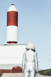 Back view man in spacesuit standing on rocky ground against striped rocket shaped antennas on sunny day - ADSF29309