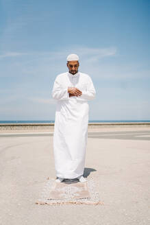 Voller Junge islamischen männlich in traditioneller weißer Kleidung stehen auf Teppich und beten gegen blauen Himmel am Strand - ADSF29318