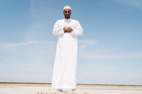 Voller Junge islamischen männlich in traditioneller weißer Kleidung stehen auf Teppich und beten gegen blauen Himmel am Strand - ADSF29319