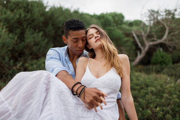 Multiracial couple of newlyweds sitting on stump of tree in woods and hugging on wedding day - ADSF29328