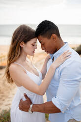 Side view of loving multiracial couple of newlyweds hugging while standing on sandy hill on background of sea on wedding day - ADSF29329