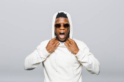 Expressive African American male in white hoodie and stylish sunglasses screaming on gray background in studio - ADSF29340