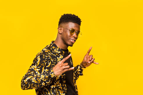 Smiling African American male in trendy outfit showing shaka gesture on yellow background in studio - ADSF29341