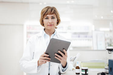 Female pharmacist with digital tablet standing in pharmacy - JOSEF05396