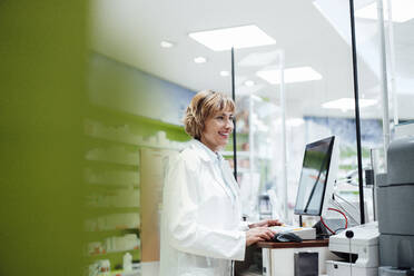 Lächelnde Apothekerin am Schreibtisch in einer Apotheke - JOSEF05403