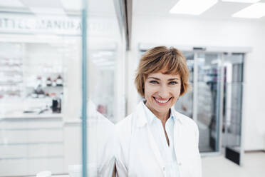 Smiling female pharmacist standing by glass wall - JOSEF05405