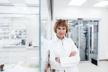 Smiling female professional with arms crossed leaning on glass wall - JOSEF05407