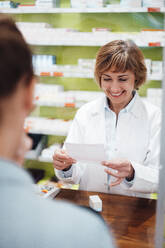 Smiling pharmacist reading prescription while standing with customer in store - JOSEF05414