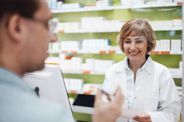 Smiling female pharmacist looking at customer checking medicine - JOSEF05416