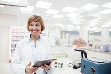 Smiling female pharmacist looking away while holding digital tablet at pharmacy - JOSEF05436