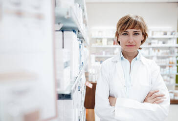 Confident female pharmacist standing by rack at medical store - JOSEF05448