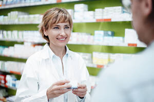 Reife Apothekerin mit Medikamenten in der Hand, während sie mit einem männlichen Kunden in einem medizinischen Geschäft spricht - JOSEF05461