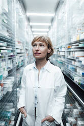 Female pharmacist with hands in pockets looking away while standing at pharmacy store - JOSEF05465