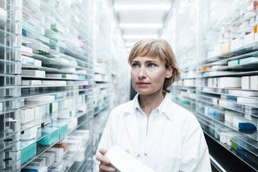 Female pharmacist with medicine looking away while standing by rack at medical store - JOSEF05466