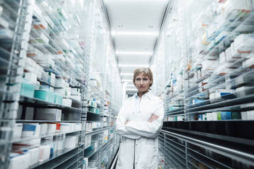 Confident female pharmacist with arms crossed standing amidst racks at medical store - JOSEF05473