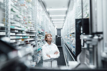 Female pharmacist with arms crossed looking at medicine in pharmacy store - JOSEF05474