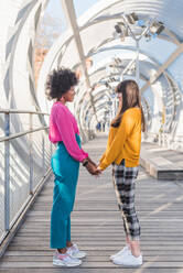 Side view of glad multiethnic LGBT couple of women holding hands while standing on bridge in city in sunny day and looking at each other - ADSF29356