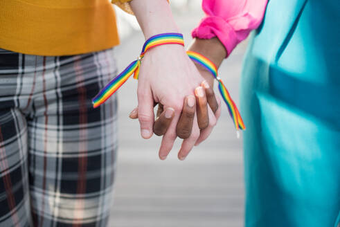 Crop unrecognizable multiethnic couple of lesbian females with LGBT rainbow bracelets holding hands in city - ADSF29358