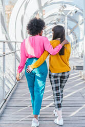 Back view of anonymous multiracial couple of homosexual females holding hands and walking along bridge in city during summer stroll - ADSF29359