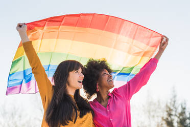 Content multiracial couple of lesbian females standing with LGBT rainbow flag in raised arms on sunny day in street - ADSF29362
