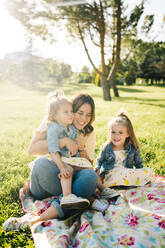 Glückliche junge Mutter mit niedlichen kleinen Töchtern in ähnlicher Kleidung genießen den sonnigen Sommertag zusammen, während sie auf einer Decke auf dem Rasen im Park sitzen - ADSF29379