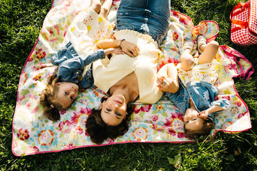 Top view of happy young woman and adorable little sisters in similar dresses lying on blanket on green grass while spending summer day together in park - ADSF29383