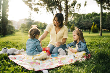 Glückliche junge Frau mit kleinen Töchtern beim Picknick auf einer grünen Wiese, während sie einen Sommertag im Park verbringen - ADSF29384