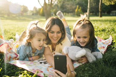 Glückliche junge Frau und niedliche kleine Töchter liegen auf der Decke und nehmen Selfie auf Smartphone, während sie Spaß zusammen auf der grünen Wiese im Sommer Park - ADSF29385