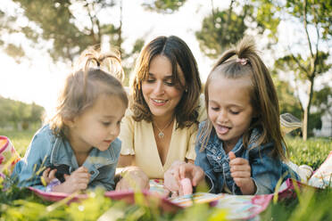 Glückliche junge Mutter mit kleinen Töchtern, die auf einer Decke liegen und ein Spiel spielen, während sie einen Sommertag im sonnigen Park verbringen - ADSF29386