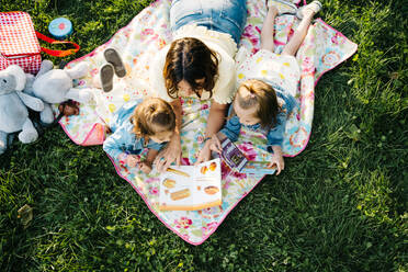 Von oben glückliche junge Mutter, die ihren kleinen Töchtern ein interessantes Buch vorliest, während sie zusammen auf einer Decke mit Spielzeug auf grünem Gras im Sommerpark liegen - ADSF29387