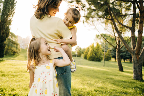 Nettes Vorschulmädchen, das lächelt und wegschaut, während es seine Mutter mit seiner kleinen Schwester an den Händen kuschelt, während eines Sommertages im grünen Park - ADSF29389