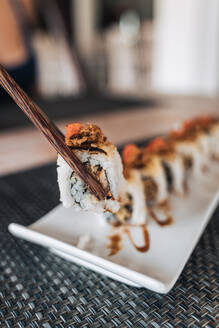 Crop person holding with chopsticks row of tasty sushi rolls with cooked rice and seafood slices on ceramic plate on table - ADSF29391