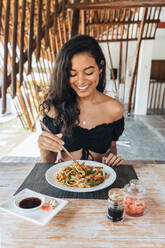 Cheerful female tourist with delicious pasta between food sticks above table with soy sauce and pickled ginger slices outdoors - ADSF29400
