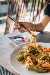 Anonymous female tourist with delicious pasta between food sticks above table with soy sauce and pickled ginger slices outdoors - ADSF29401