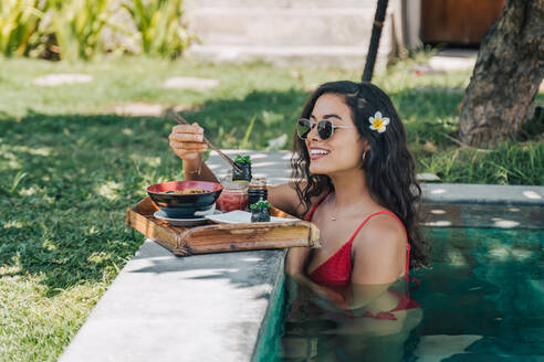 Seitenansicht einer fröhlichen weiblichen Reisenden in Badekleidung am Pool mit lecker gekochten asiatischen Nudeln zwischen Stäbchen im Sonnenlicht - ADSF29404