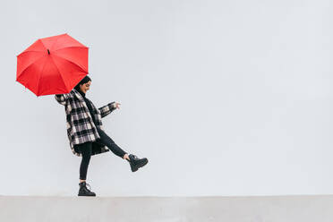 Happy young female with red umbrella walking and balancing on border against gray wall on rainy day on street - ADSF29410